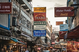 A bustling urban street filled with various signs in multiple languages, indicating different businesses such as restaurants and car parks. The scene is characterized by a mix of cultural influences and is crowded with overhead wires and tightly packed buildings.