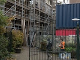 A construction site with scaffolding on a building, accompanied by two workers in safety gear walking nearby. The scene includes an urban setting with a metal fence in the foreground and a shipping container, along with graffiti and vegetation such as bushes and potted plants.