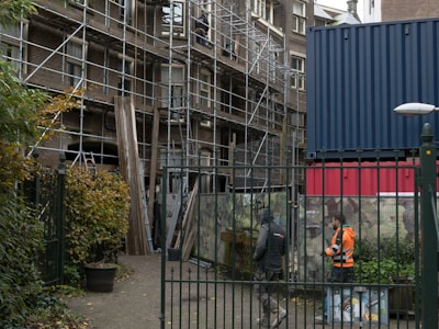 A construction site with scaffolding on a building, accompanied by two workers in safety gear walking nearby. The scene includes an urban setting with a metal fence in the foreground and a shipping container, along with graffiti and vegetation such as bushes and potted plants.