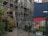 A construction site with scaffolding on a building, accompanied by two workers in safety gear walking nearby. The scene includes an urban setting with a metal fence in the foreground and a shipping container, along with graffiti and vegetation such as bushes and potted plants.