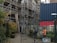 A construction site with scaffolding on a building, accompanied by two workers in safety gear walking nearby. The scene includes an urban setting with a metal fence in the foreground and a shipping container, along with graffiti and vegetation such as bushes and potted plants.