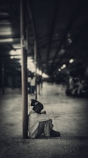 A person sits alone on the ground in a dark, empty space. The figure leans against a pole with their head resting on their knees, conveying a sense of solitude. The surrounding area is dimly lit, with blurred lights in the background creating a feeling of distance and isolation.