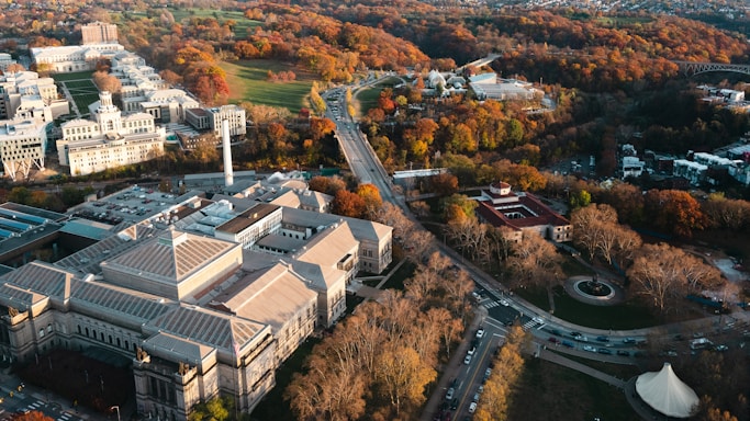 Aerial view of a cityscape featuring a large museum-like building with a triangular roof, surrounded by a mix of modern and historic architectures. The landscape is dotted with trees showcasing vibrant autumn colors. Roads and cars are visible, creating a sense of urban life, while a park area with open green spaces and walkways adds natural beauty.