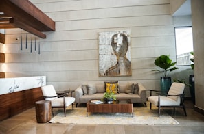 A contemporary and stylish lobby lounge with a modern art piece on the wall. The seating area includes a grey sofa, two beige armchairs, and a wooden coffee table with a green plant. The space is complemented by wooden accents, a marble wall feature, and several potted plants, creating a welcoming and sophisticated ambiance.
