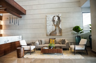 A contemporary and stylish lobby lounge with a modern art piece on the wall. The seating area includes a grey sofa, two beige armchairs, and a wooden coffee table with a green plant. The space is complemented by wooden accents, a marble wall feature, and several potted plants, creating a welcoming and sophisticated ambiance.