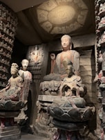 A group of intricately carved Buddhist statues is displayed in an ancient stone grotto. The tallest statue is of a serene Buddha with a delicate, peaceful expression, seated on a lotus pedestal. Smaller figures are situated around the main Buddha, each intricately detailed with traditional garments and headpieces. The overall setting is dim, with a soft spotlight highlighting the expressive features and detailed carvings on the statues.