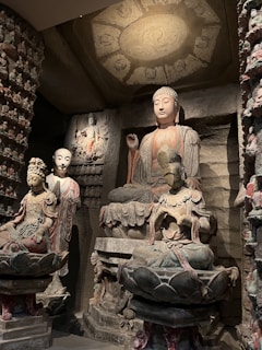 A group of intricately carved Buddhist statues is displayed in an ancient stone grotto. The tallest statue is of a serene Buddha with a delicate, peaceful expression, seated on a lotus pedestal. Smaller figures are situated around the main Buddha, each intricately detailed with traditional garments and headpieces. The overall setting is dim, with a soft spotlight highlighting the expressive features and detailed carvings on the statues.