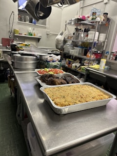 A stainless steel kitchen table is filled with trays of prepared food, including a large tray of rice and roasted vegetables. In the background, shelves hold various kitchen supplies such as spices, dishes, and cleaning products. Hanging utensils are visible above the table.