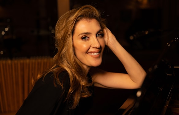 A woman with long, wavy hair is smiling warmly while seated by a piano. The ambiance is dimly lit, creating a cozy and intimate atmosphere.