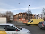 A modern building with a unique design featuring wooden diagonal panels on its facade. It has signage that appears to include company names. Several vehicles, including a Yandex Taxi and a yellow van, are parked nearby. There are trees without leaves and a bird flying above.