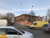 A modern building with a unique design featuring wooden diagonal panels on its facade. It has signage that appears to include company names. Several vehicles, including a Yandex Taxi and a yellow van, are parked nearby. There are trees without leaves and a bird flying above.