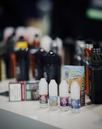 Small bottles of flavored e-liquid and vape juice are arranged in the foreground with larger vape devices and packaging blurred in the background. The bottles display colorful labeling and branding.