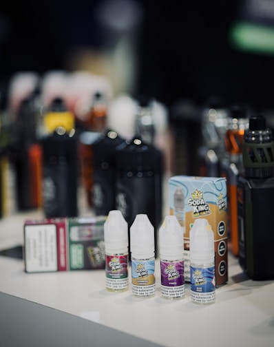 Small bottles of flavored e-liquid and vape juice are arranged in the foreground with larger vape devices and packaging blurred in the background. The bottles display colorful labeling and branding.