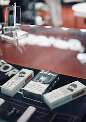 A display case containing a variety of boxed electronic cigarette products and accessories. The background is slightly blurred with a dominant red color. The boxes have text indicating nicotine content and warnings about nicotine being addictive. A device is placed upright in a transparent stand.