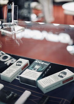 A display case containing a variety of boxed electronic cigarette products and accessories. The background is slightly blurred with a dominant red color. The boxes have text indicating nicotine content and warnings about nicotine being addictive. A device is placed upright in a transparent stand.