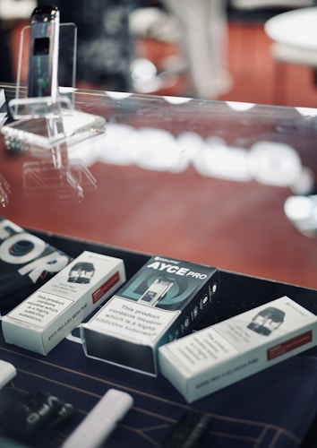 A display case containing a variety of boxed electronic cigarette products and accessories. The background is slightly blurred with a dominant red color. The boxes have text indicating nicotine content and warnings about nicotine being addictive. A device is placed upright in a transparent stand.