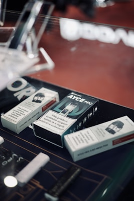 Several boxes of vaping products are displayed on a countertop. The boxes contain warnings about nicotine being a highly addictive substance. The setting appears to be a retail environment, possibly a vape shop.