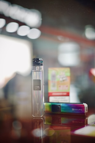 A sleek electronic device with a metallic, iridescent surface standing next to a colorful product packaging box. The device is reflective and appears modern, with branding visible on its body. The background is softly blurred, adding depth to the image.