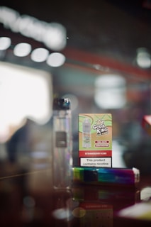 A close-up of a vaping product display featuring a cardboard box labeled 'Soda King Bar Salts' with the flavor 'Strawberry Kiwi' and a nicotine content warning. Beside it is a rainbow-colored vape pen. The background is blurred, giving a focus on the products.