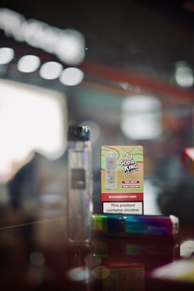 A close-up of a vaping product display featuring a cardboard box labeled 'Soda King Bar Salts' with the flavor 'Strawberry Kiwi' and a nicotine content warning. Beside it is a rainbow-colored vape pen. The background is blurred, giving a focus on the products.