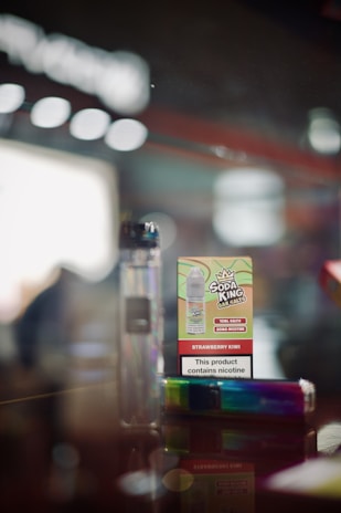 A close-up of a vaping product display featuring a cardboard box labeled 'Soda King Bar Salts' with the flavor 'Strawberry Kiwi' and a nicotine content warning. Beside it is a rainbow-colored vape pen. The background is blurred, giving a focus on the products.