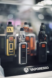 Several electronic vaping devices are displayed in a neatly organized manner. The devices have different color schemes including yellow and black, silver and black, orange and black, and all black. They are placed on a stand with a logo that reads 'Vaporesso'. The background is slightly out of focus with soft lighting and bokeh effects, giving a sleek and modern appearance.
