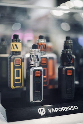 Several electronic vaping devices are displayed in a neatly organized manner. The devices have different color schemes including yellow and black, silver and black, orange and black, and all black. They are placed on a stand with a logo that reads 'Vaporesso'. The background is slightly out of focus with soft lighting and bokeh effects, giving a sleek and modern appearance.