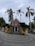 A red brick church with a prominent cross at the top is surrounded by tall palm trees and a wrought iron gate. The entrance path is paved with concentric circular patterns, and the sky appears overcast.