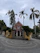 A red brick church with a prominent cross at the top is surrounded by tall palm trees and a wrought iron gate. The entrance path is paved with concentric circular patterns, and the sky appears overcast.