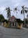 A red brick church with a prominent cross at the top is surrounded by tall palm trees and a wrought iron gate. The entrance path is paved with concentric circular patterns, and the sky appears overcast.