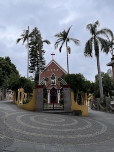 A red brick church with a prominent cross at the top is surrounded by tall palm trees and a wrought iron gate. The entrance path is paved with concentric circular patterns, and the sky appears overcast.