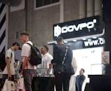 Several people are gathered in what appears to be a trade show or convention setting. They are standing near a display counter with products and packaging. The background features signage with the text 'DOVPO' and a website address. One person is holding shopping bags, and others appear to have casual conversations, giving a social atmosphere.