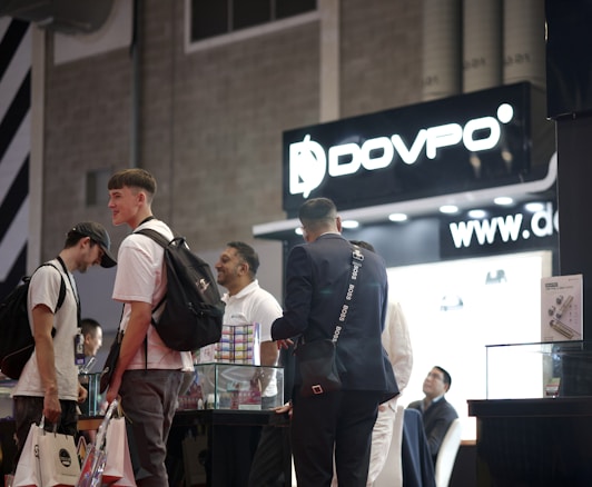 Several people are gathered in what appears to be a trade show or convention setting. They are standing near a display counter with products and packaging. The background features signage with the text 'DOVPO' and a website address. One person is holding shopping bags, and others appear to have casual conversations, giving a social atmosphere.