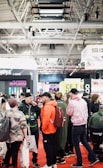 A bustling convention or trade show setting with numerous people gathered indoors. The ceiling is industrial with visible beams and lighting fixtures. Attendees are casually dressed, some carrying bags with branding. Booths line the background, featuring various advertisements and displays. The atmosphere suggests a lively and social environment.