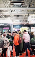 A bustling convention or trade show setting with numerous people gathered indoors. The ceiling is industrial with visible beams and lighting fixtures. Attendees are casually dressed, some carrying bags with branding. Booths line the background, featuring various advertisements and displays. The atmosphere suggests a lively and social environment.