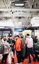 A bustling convention or trade show setting with numerous people gathered indoors. The ceiling is industrial with visible beams and lighting fixtures. Attendees are casually dressed, some carrying bags with branding. Booths line the background, featuring various advertisements and displays. The atmosphere suggests a lively and social environment.