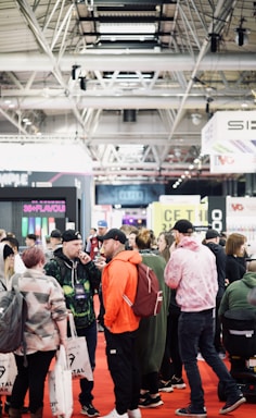 A bustling convention or trade show setting with numerous people gathered indoors. The ceiling is industrial with visible beams and lighting fixtures. Attendees are casually dressed, some carrying bags with branding. Booths line the background, featuring various advertisements and displays. The atmosphere suggests a lively and social environment.