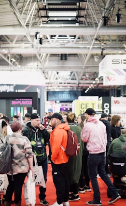 A bustling convention or trade show setting with numerous people gathered indoors. The ceiling is industrial with visible beams and lighting fixtures. Attendees are casually dressed, some carrying bags with branding. Booths line the background, featuring various advertisements and displays. The atmosphere suggests a lively and social environment.