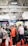 A bustling convention or trade show setting with numerous people gathered indoors. The ceiling is industrial with visible beams and lighting fixtures. Attendees are casually dressed, some carrying bags with branding. Booths line the background, featuring various advertisements and displays. The atmosphere suggests a lively and social environment.