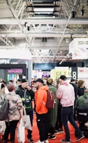 A bustling convention or trade show setting with numerous people gathered indoors. The ceiling is industrial with visible beams and lighting fixtures. Attendees are casually dressed, some carrying bags with branding. Booths line the background, featuring various advertisements and displays. The atmosphere suggests a lively and social environment.