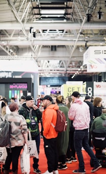 A bustling convention or trade show setting with numerous people gathered indoors. The ceiling is industrial with visible beams and lighting fixtures. Attendees are casually dressed, some carrying bags with branding. Booths line the background, featuring various advertisements and displays. The atmosphere suggests a lively and social environment.