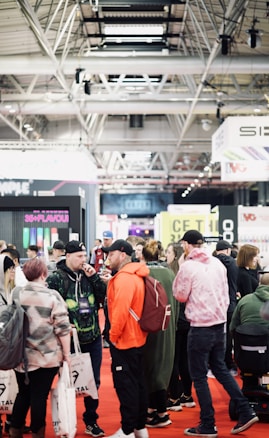 A bustling convention or trade show setting with numerous people gathered indoors. The ceiling is industrial with visible beams and lighting fixtures. Attendees are casually dressed, some carrying bags with branding. Booths line the background, featuring various advertisements and displays. The atmosphere suggests a lively and social environment.