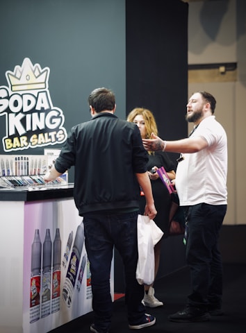 A group of people stands next to a booth displaying a variety of vape products. The booth is branded with 'Soda King Bar Salts', and there are several vaping liquid bottles showcased with different flavors. One person appears to be explaining something to another, while a third person holds a bag and listens.