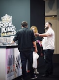 A group of people stands next to a booth displaying a variety of vape products. The booth is branded with 'Soda King Bar Salts', and there are several vaping liquid bottles showcased with different flavors. One person appears to be explaining something to another, while a third person holds a bag and listens.