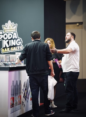 A group of people stands next to a booth displaying a variety of vape products. The booth is branded with 'Soda King Bar Salts', and there are several vaping liquid bottles showcased with different flavors. One person appears to be explaining something to another, while a third person holds a bag and listens.
