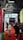 A busy convention or trade show setting with a crowd of people walking along a red carpet between rows of exhibition booths. Overhead, a large advertisement for 'AYCE PRO' vape products is displayed. The hall's architecture features visible metal beams and a high ceiling.