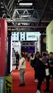 A busy convention or trade show setting with a crowd of people walking along a red carpet between rows of exhibition booths. Overhead, a large advertisement for 'AYCE PRO' vape products is displayed. The hall's architecture features visible metal beams and a high ceiling.