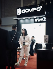 A trade show or exhibition setting featuring a booth with a large sign displaying the brand 'DOVPO'. A woman in a white outfit is interacting with a man in a suit, while other people are seated in the background. There is a product display case and marketing materials visible.