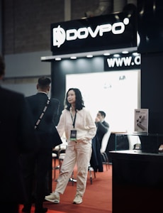 A trade show or exhibition setting featuring a booth with a large sign displaying the brand 'DOVPO'. A woman in a white outfit is interacting with a man in a suit, while other people are seated in the background. There is a product display case and marketing materials visible.