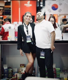 A man and a woman are posing together at an event, both wearing exhibitor badges. The man is exhaling vapor from an e-cigarette, creating a cloud of smoke. The background features signs and a flag, suggesting a diverse or international setting.